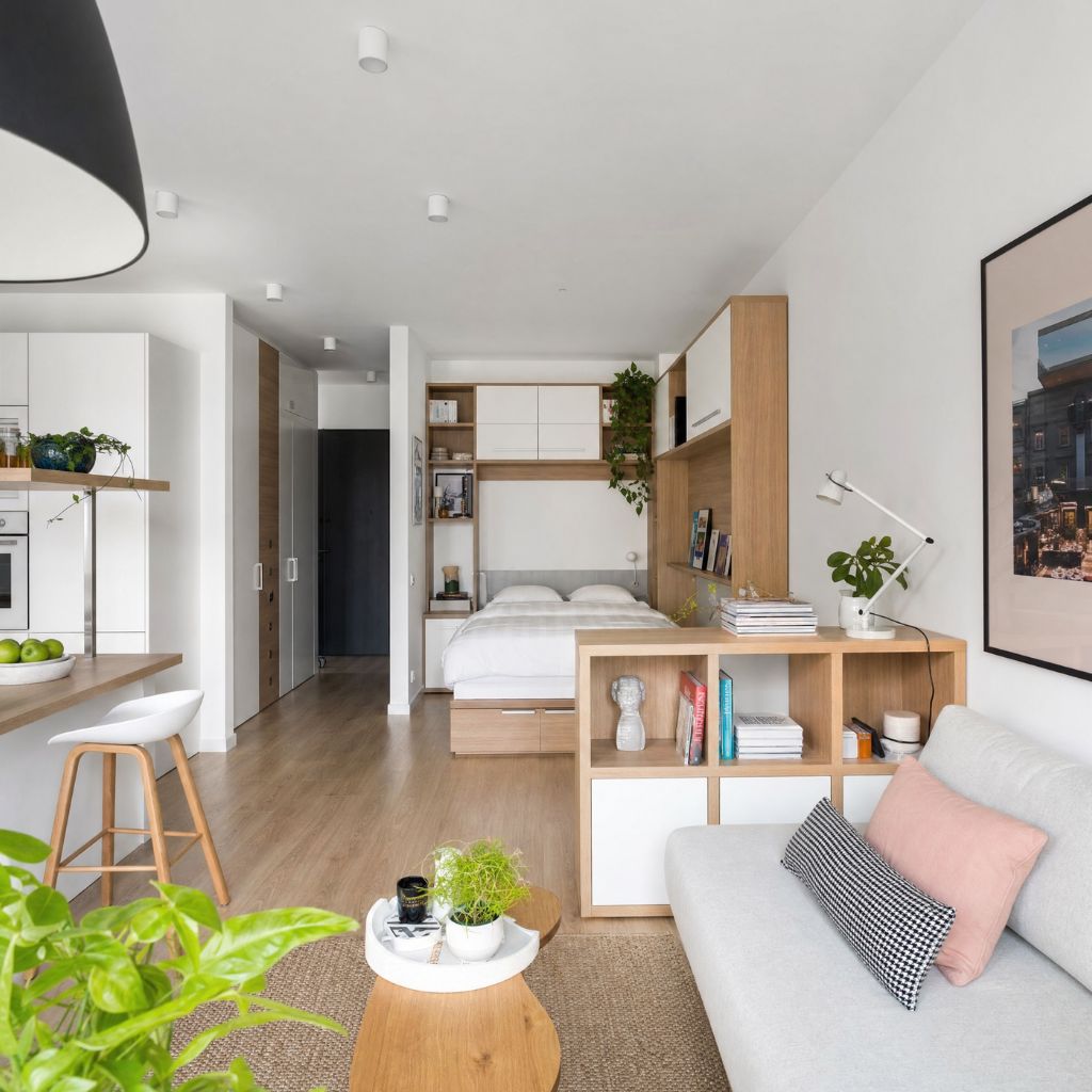 Open-plan studio with a living area, dining island, kitchenette, and bed at the back; light wood furniture and greenery throughout.
