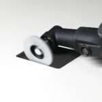 Close-up of a power sander with a spinning circular sanding disk contacting a black square sheet of material at an angle.