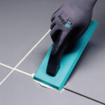 Close-up of a gloved hand using a teal grout float to apply white grout between gray tiles in a flooring project.