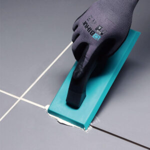 Close-up of a gloved hand using a teal grout float to apply white grout between gray tiles in a flooring project.