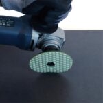 Gloved hand holds a rotary power tool with a green honeycomb-pattern polishing disc attached to the spindle surface.
