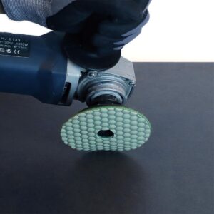 Gloved hand holds a rotary power tool with a green honeycomb-pattern polishing disc attached to the spindle surface.