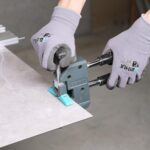 Person wearing gray gloves using a tile edge trimming tool to cut a light gray marble tile on a workbench