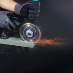 Worker wearing gloves uses a handheld grinder to cut metal, sparks flying from the cut edge.