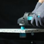 Gloved hand operating a handheld power tool with a turquoise switch on a granite work surface.