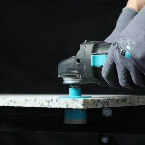 Gloved hand operating a handheld power tool with a turquoise switch on a granite work surface.